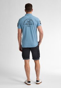 Ljusblå pikétröja med svart tryckt text på ryggen, ihop med mörka cargoshorts och vita skor. Snyggt tyg, avslappnad design.