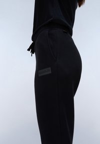 Zwarte joggingsbroek met een elastische tailleband en aantrekkoord, voorzien van zijzakken en een gestructureerde logopatch op de linker dij.