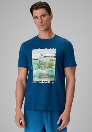 Mężczyzna w niebieskiej koszulce z nadrukiem plaży z palmami, falami oceanu i kamperem, stojący na jednolitym szarym tle.