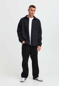 Black button-up shirt with a textured fabric, front pocket, worn over a white t-shirt; paired with black pants and white sneakers.