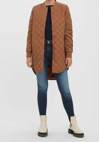 Veste longue matelassée marron avec un col rond, des boutons-pression et des poignets côtelés, portée sur un haut bleu marine et un jean skinny bleu, assortie à des bottes beiges.