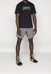 Hombre con camiseta negra de Venum, pantalones cortos deportivos grises y zapatillas altas deportivas en negro y beige, de pie contra un fondo blanco plano.