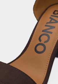 Κοντινό πλάνο καφέ πέδιλου με τακούνι, με ταμπά εσωτερική σόλα και την επωνυμία "BIANCO" τυπωμένη με μαύρα γράμματα.