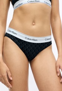 Czarne majtki bikini z subtelnym wzorem geometrycznym, z białym elastycznym pasem na biodrach oznaczonym logo "Calvin Klein".