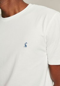 Valge puuvillane T-särk lühikeste varrukatega, mille vasakul rinnal on väike sinine jänese tikand. Sile tekstuur ja klassikaline ümar kaelus.