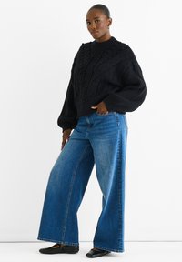 Maglione nero a maglia con collo alto, abbinato a jeans in denim blu a gamba larga. L'outfit presenta una vestibilità comoda e dettagli testurizzati.