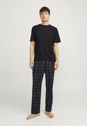Zwarte korte mouwen t-shirt gecombineerd met marineblauwe geruite pyjamabroek met witte en blauwe lijnen. Model staat op blote voeten.