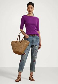 Γυναίκα με μοβ εφαρμοστό μπλουζάκι, τζιν παντελόνι κατευθυνόμενο που είναι μπαγιάτικο από μπογιές, καφέ σανδάλια και κρατάει μια μεγάλη πλεκτή τσάντα χειρός.