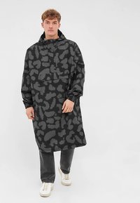 Zwarte pullover jas met grijs abstract patroon, voorzien van een hoge hals, lange mouwen en een borstzak; gedragen over donkere broek en schoenen.