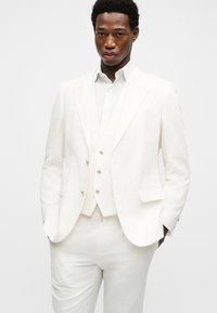 Άνδρας που φοράει ένα λευκό κοστούμι τριών τεμαχίων με σακάκι, γιλέκο και παντελόνι, στέκεται με τα χέρια στις τσέπες μπροστά σε απλό φόντο.