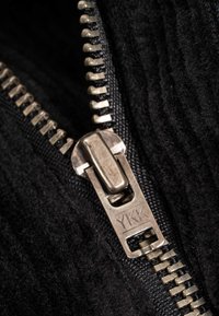 Black textured fabric with a silver metal zipper. Zipper pull features "YKK" branding. Close-up view highlights stitching and fabric detail.