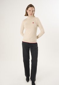 Maglione beige a collo alto con un piccolo dettaglio a forma di cuore rosso, abbinato a jeans neri a gamba dritta e stivaletti neri con tacco. Design semplice.