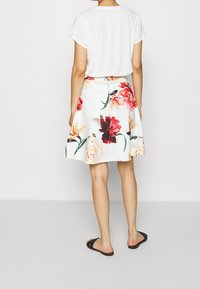 Žena v bílém triku s krátkým rukávem a bílou sukni s velkými květinovými vzory v červené, oranžové a žluté barvě, doplněná černými pantofli.