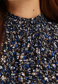 Blus med hög krage och smockad krage i svart tyg som har ett intrikat blommönster i blått, vitt och brunt.