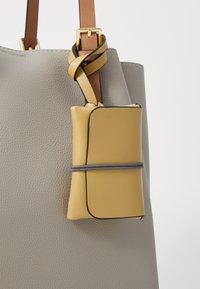 Pelēka ādas soma ar teksturētu virsmu, ar dzeltenu ādas maisiņu, kas piestiprināts ar brūnu siksnu un pelēku elastīgu joslu.