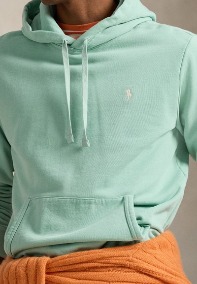 Světlá mátově zelená mikina z měkké látky, s předním kapsou a šňůrkami, s malým bílým logem na hrudi. Pod tím oranžová pletená vrstva.