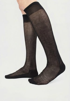 Voeten met doorschijnende zwarte kniekousen met versterkte tenen en boorden, staand tegen een effen witte achtergrond.