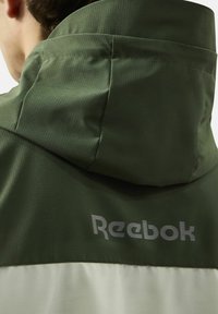 Groene hoodie met een gestructureerde afwerking, voorzien van een witte onderzijde en een reflecterend 'Reebok' logo op de achterkant.