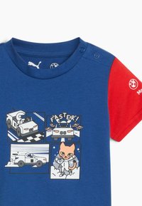 Kreklu ar īsām piedurknēm zilā krāsā ar sarkano piedurkni. Tās dizainā ir zīmējumi ar multfilmu automašīnu un kaķi. Izgatavots no kokvilnas, ar apaļu kakla izgriezumu un pogām pie pleca.