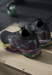 Paire de chaussures de sport noires avec des semelles intermédiaires colorées et à motifs, posées sur une surface en bois dans une salle de sport.