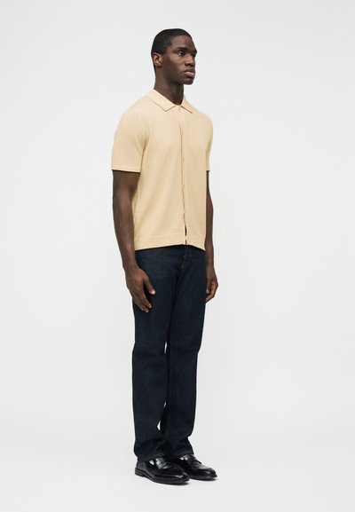 Άνδρας στέκεται στραμμένος προς τα δεξιά φορώντας μπεζ πουκάμισο με κοντά μανίκια και κουμπιά, σκουρόχρωμο μπλε τζιν και μαύρα παπούτσια επίσημα, με φόντο λευκό.