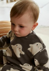 Grijze katoenen rompertje met een spookpatroon in crème, voorzien van geprinte gezichten en het woord "BABY" op de mouw. Zachte textuur, casual ontwerp.