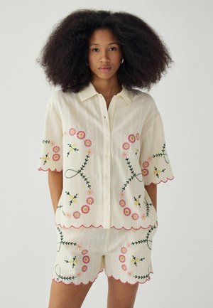 Kobieta ubrana w kremową koszulę na guziki z krótkim rękawem oraz pasujące krótkie spodenki ozdobione różowo-żółtą kwiatową wyszywanką i falistymi brzegami.