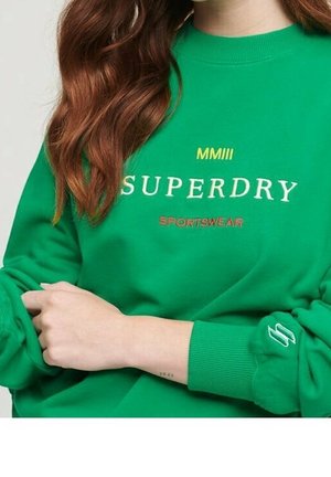 Zielona bluza z napisem "MMIII SUPERDRY SPORTSWEAR", długie czerwone włosy oraz podwinięte rękawy z wyhaftowanym logo.