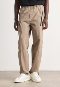 Beige cargo pants with an elastic waistband, side zip pockets, and stitched detailing. Material appears lightweight and textured.
