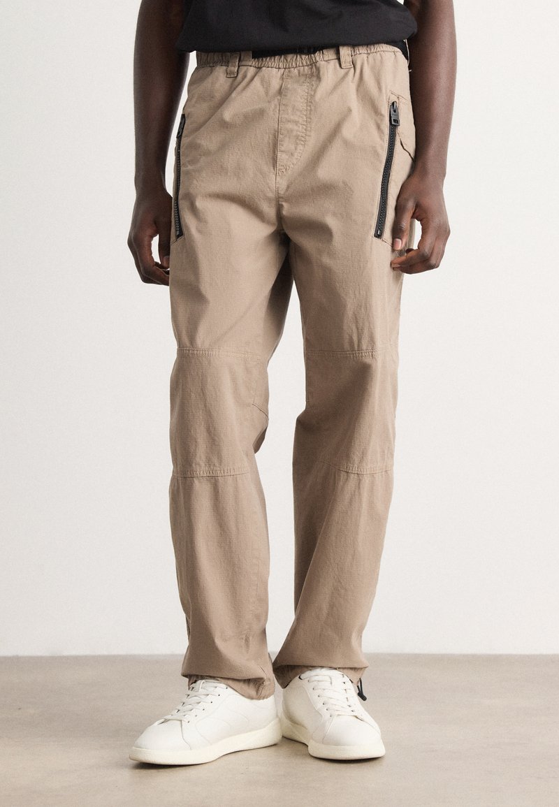 Beige cargo pants with an elastic waistband, side zip pockets, and stitched detailing. Material appears lightweight and textured.