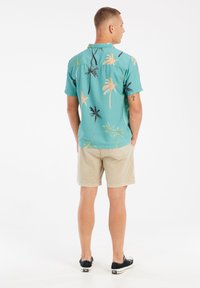 Korte mouw, turkooise overhemd met knopen en een palmboomprint in perzik en zwart, gecombineerd met beige shorts en zwarte sneakers.
