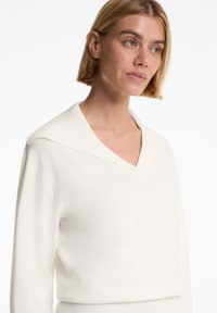 Witte gebreide trui met een diepe V-hals en een brede kraag, met een zachte textuur en lange mouwen. Eenvoudig, strak ontwerp zonder patronen.