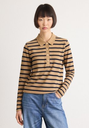 Kvinne med kort svart hår iført en tan og svart stripet langermet polo-skjorte og blå jeans, stående med den ene hånden i lommen.