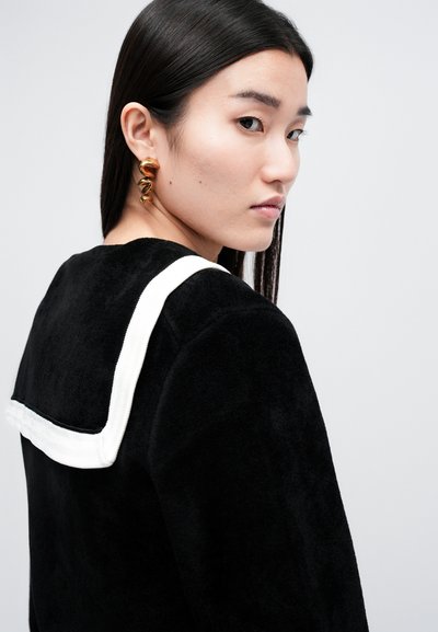 Schwarzes, flauschiges Oberteil mit einem kontrastierenden weißen Trägerdetail, das einen strukturierten Stoff und ein schlichtes, modernes Design zeigt. Goldene, gedrehte Ohrringe vervollständigen den Look.