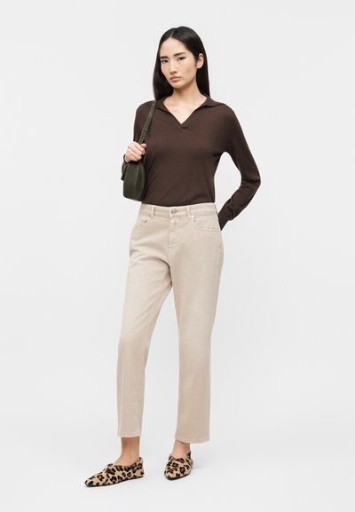 Femme aux longs cheveux noirs portant un haut marron à manches longues, un pantalon beige, des ballerines à imprimé léopard et un sac à bandoulière vert.