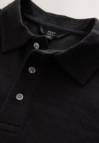Polo noir en tissu texturé, avec un col, une patte de boutonnage à trois boutons et des boutons argentés. L'étiquette indique "NEXT REGULAR FIT".