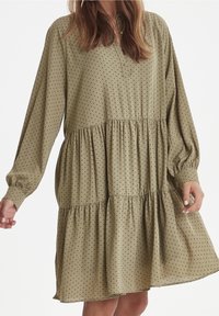 Olivově zelené šaty s černými puntíky, dlouhými rukávy, knoflíkovým límcem a vrstveným volánkovým designem v pase. Měkká textura látky.