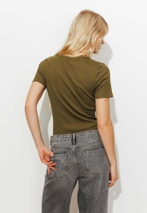 Femme blonde vue de dos, portant un haut à manches courtes vert olive et un jean taille haute gris, la main posée sur la poche arrière.