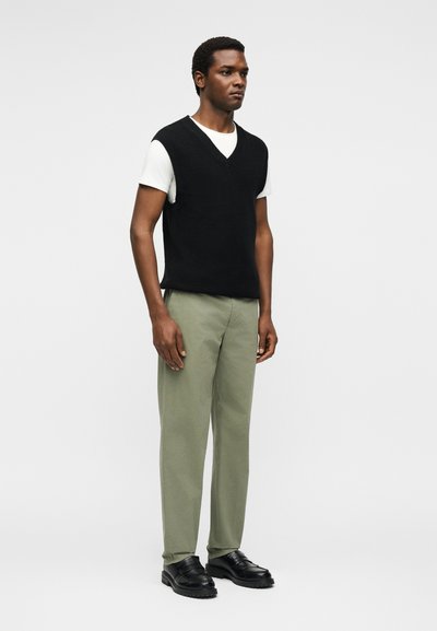 Άνδρας στέκεται φορώντας μαύρο αμάνικο πουλόβερ με V-λαιμόκοψη πάνω από λευκό μπλουζάκι, χακί παντελόνι και μαύρα μοκασίνια μπροστά σε απλό ανοιχτό φόντο.