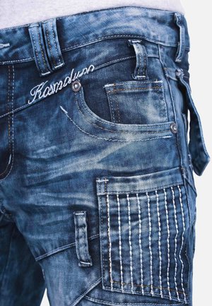 Denim jeans in dark blue with faded accents, featuring multiple pockets, intricate stitching on one pocket, and a visible brand logo.