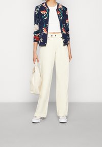 Blomsterprydd bomberjacka i marinblått, vit crop top, vida krämfärgade byxor och vita sneakers som håller en krämfärgad väska.