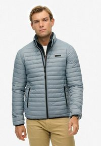 Mężczyzna w jasnoszarej pikowanej kurtce, noszący czarną bluzę na zamek i beżowe spodnie, stojący na tle białego tła.