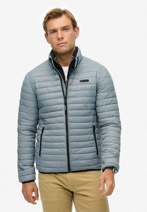 Man wearing light gray quilted jacket over black zip-up layer and beige pants, standing against a plain white background.