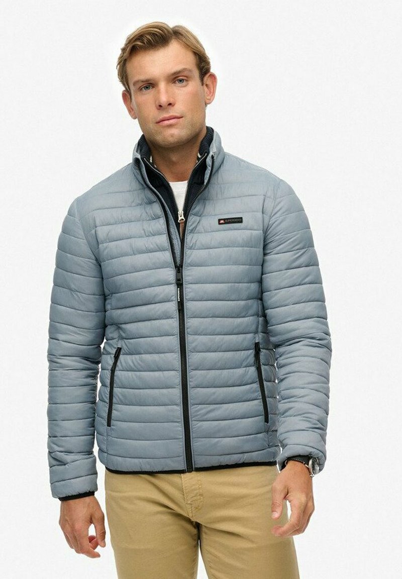 Mężczyzna w jasnoszarej pikowanej kurtce, noszący czarną bluzę na zamek i beżowe spodnie, stojący na tle białego tła.