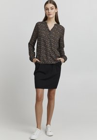 Svart bluse med blomsterdesign, lange ermer og V-hals, kombinert med et svart skjørt. Antrekket kompletteres med hvite sneakers for et avslappet utseende.