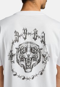 Witte T-shirt met een zwart grafisch ontwerp van een brullende tijger, omringd door prikkeldraad, rozen en yin-yang symbolen.