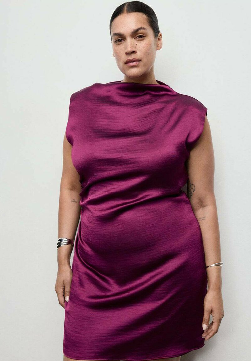 Mujer con pelo oscuro recogido que lleva un vestido sin mangas de un brillante color magenta y pulseras de plata en ambas muñecas, de pie contra un fondo claro y sencillo.