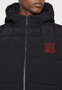 Schwarze gepolsterte Jacke mit Reißverschluss und Kapuze. Verfügt über das rot gestickte "HUGO"-Logo auf der Brust und eine gesteppten Struktur.