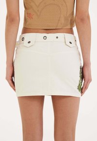 Gonna mini in denim bianco con una silhouette aderente, caratterizzata da un design grafico verde sul lato destro e dettagli in metallo argentato con bottoni e chiusure a scatto.