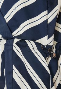 Tessuto blu navy con strisce diagonali bianche e dettaglio con cordino. Include un elemento logo e una texture liscia. Vista ravvicinata della sezione del capo.